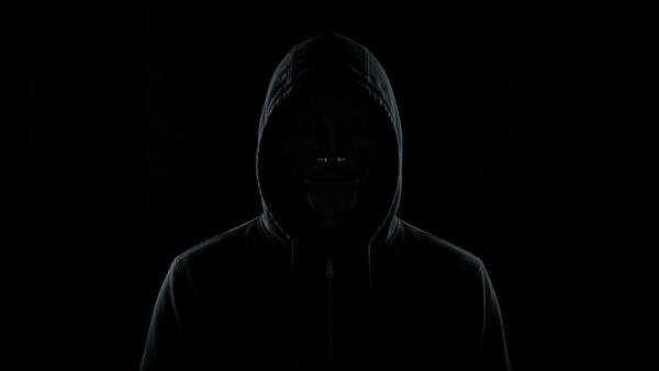 The picture shows a hacker in black clothing with weird eyes in a very dark room